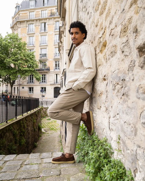 Man leaning against a Parisian stone wall in an urban setting, wearing suede barefoot derby shoes