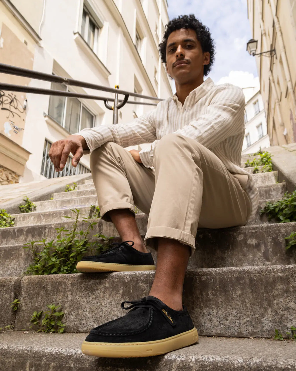 Man sitting on steps wearing a white shirt, beige pants, and black barefoot derby shoes.
