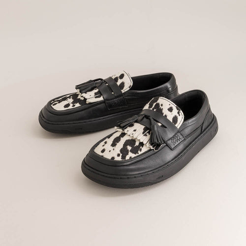 Pair of black barefoot loafers with white cow print soles on a light gray background