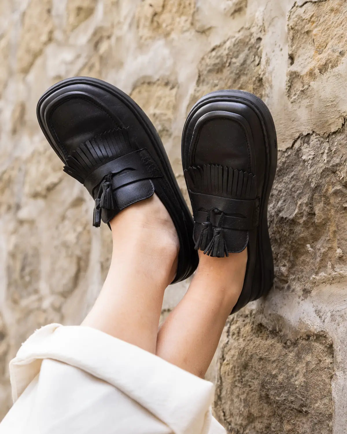 Black loafers with tassels worn by a person against a stone wall.