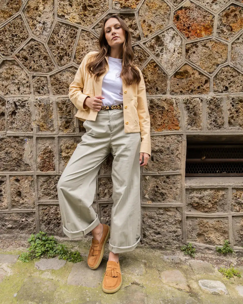 Woman in a yellow jacket and light pants standing against a stone wall, wearing suede barefoot loafers