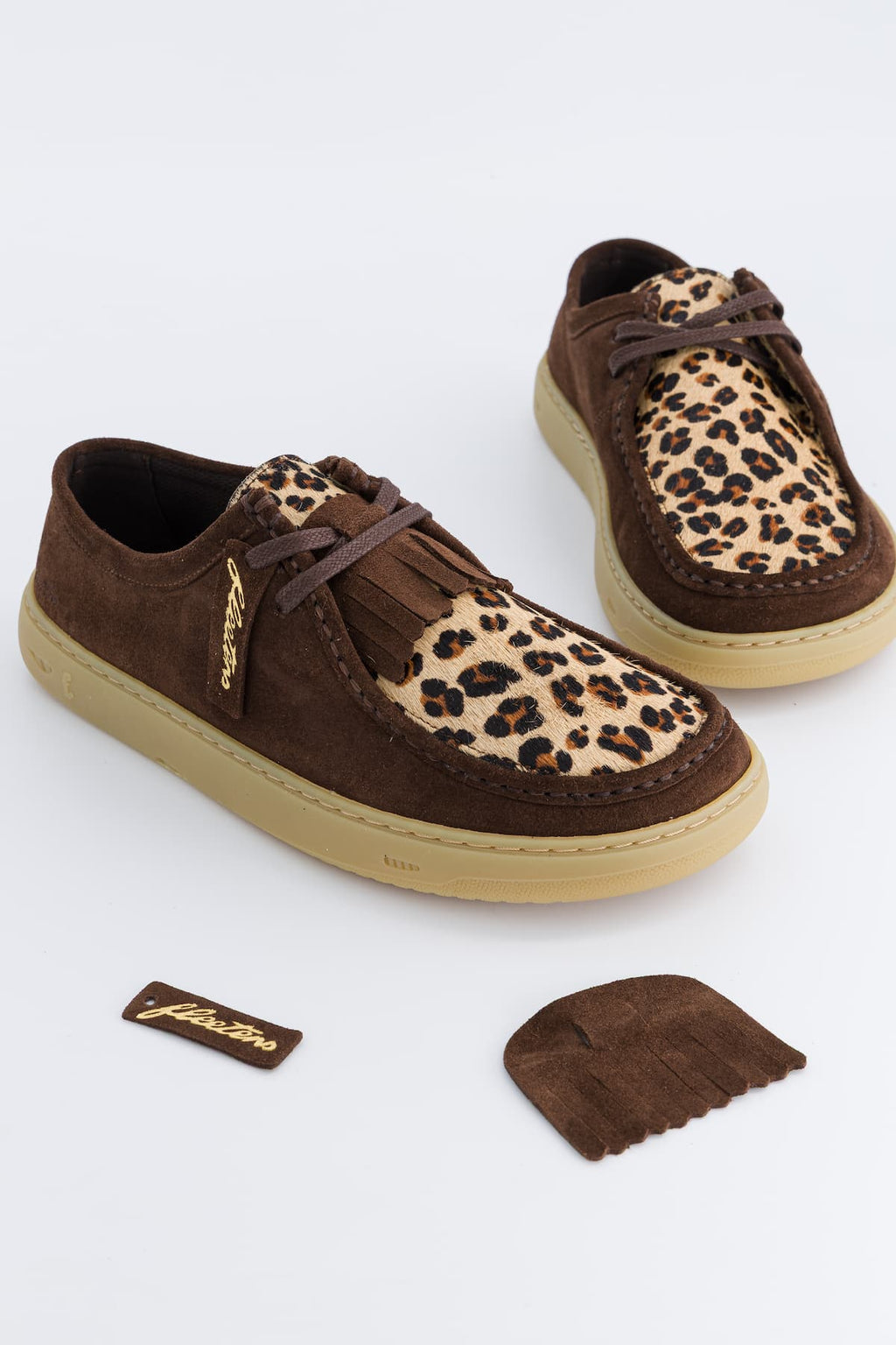 A pair of brown suede and leopard-print barefoot sneakers with tassel laces, displayed on a clean white background with a small brand tag and a detached tassel piece below.