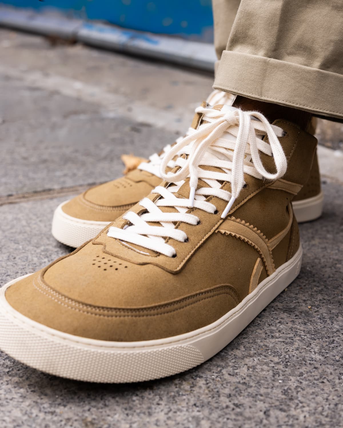A close-up shot of a person wearing tan suede high-top barefoot sneakers with white laces and off-white rubber soles, paired with khaki pants on a gray concrete ground.