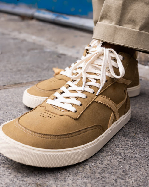 A close-up shot of a person wearing tan suede high-top barefoot sneakers with white laces and off-white rubber soles, paired with khaki pants on a gray concrete ground.