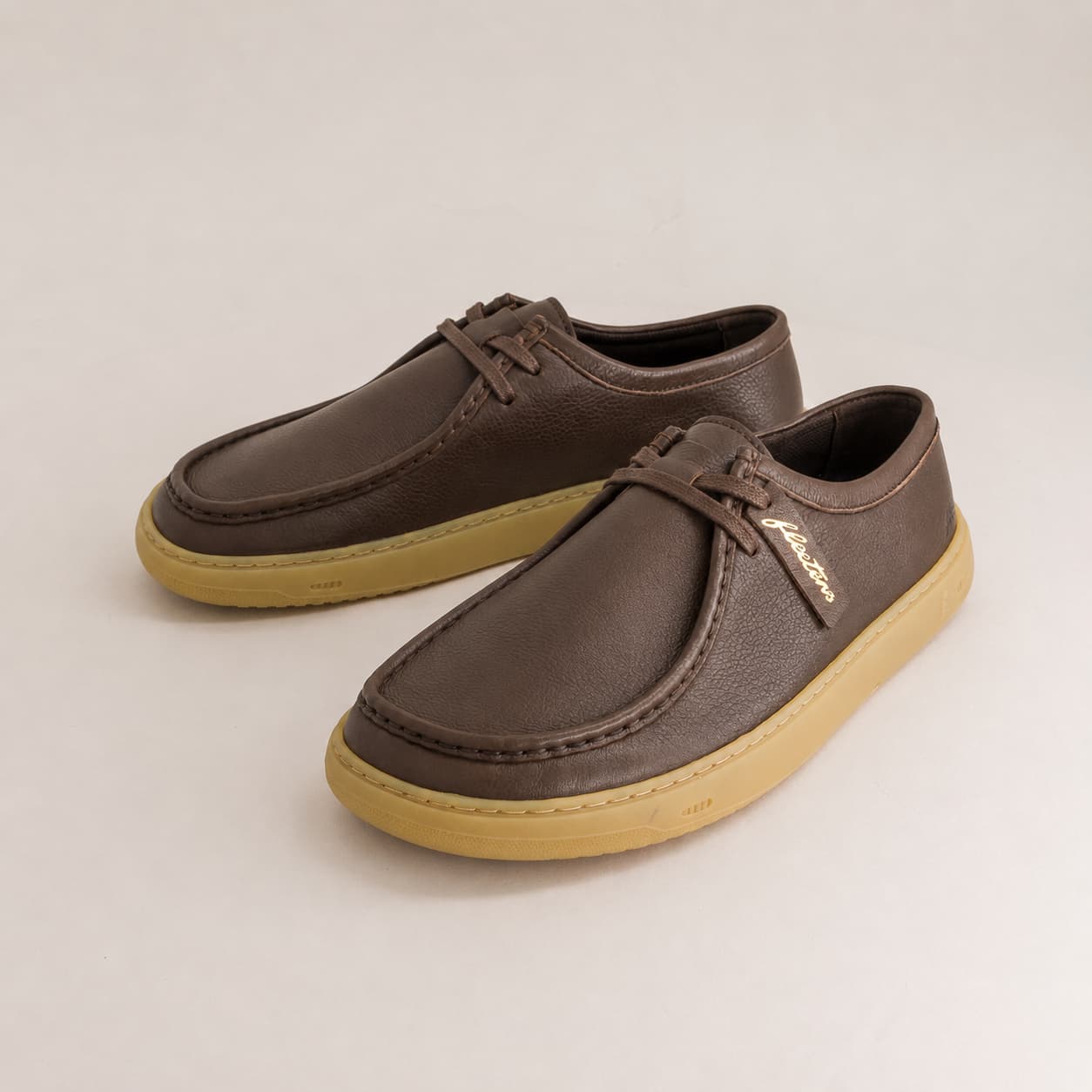 A pair of dark brown leather lace-up shoes with tan soles, displayed on a light gray background. A small gold brand tag is attached to the laces.