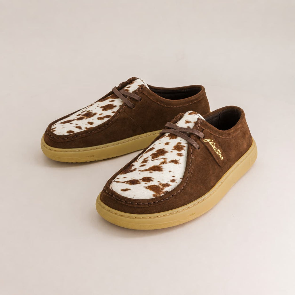 Une paire de chaussures en suédine marron à empeigne motif vache blanc et semelles beiges, sur fond beige clair.