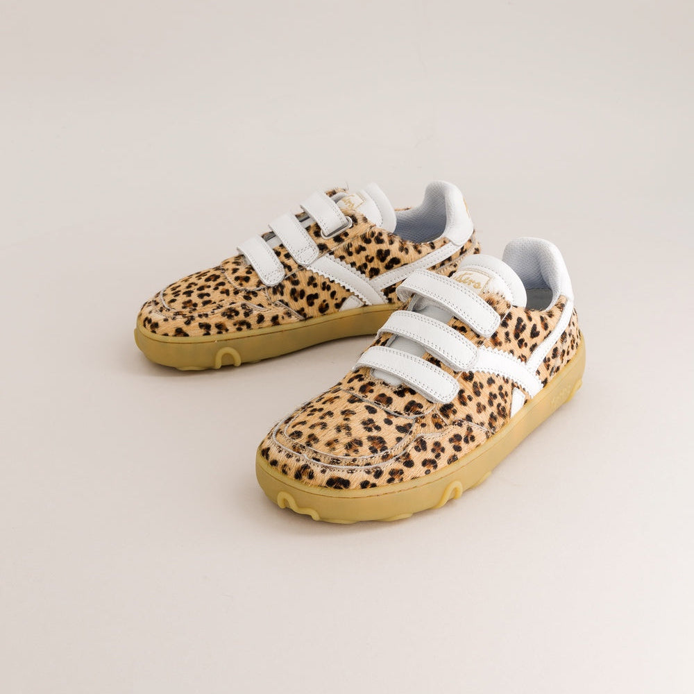 A pair of children's barefoot sneakers featuring a brown and black leopard print pattern, white hook-and-loop strap closures, and gum rubber soles, displayed on a light beige studio background.