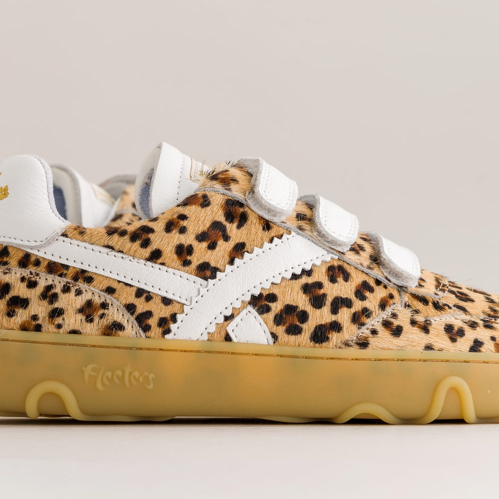 A close-up side view of a low-top sneaker featuring a beige and black leopard print pattern, white side stripes and velcro-style laces, and a translucent yellow gum sole.