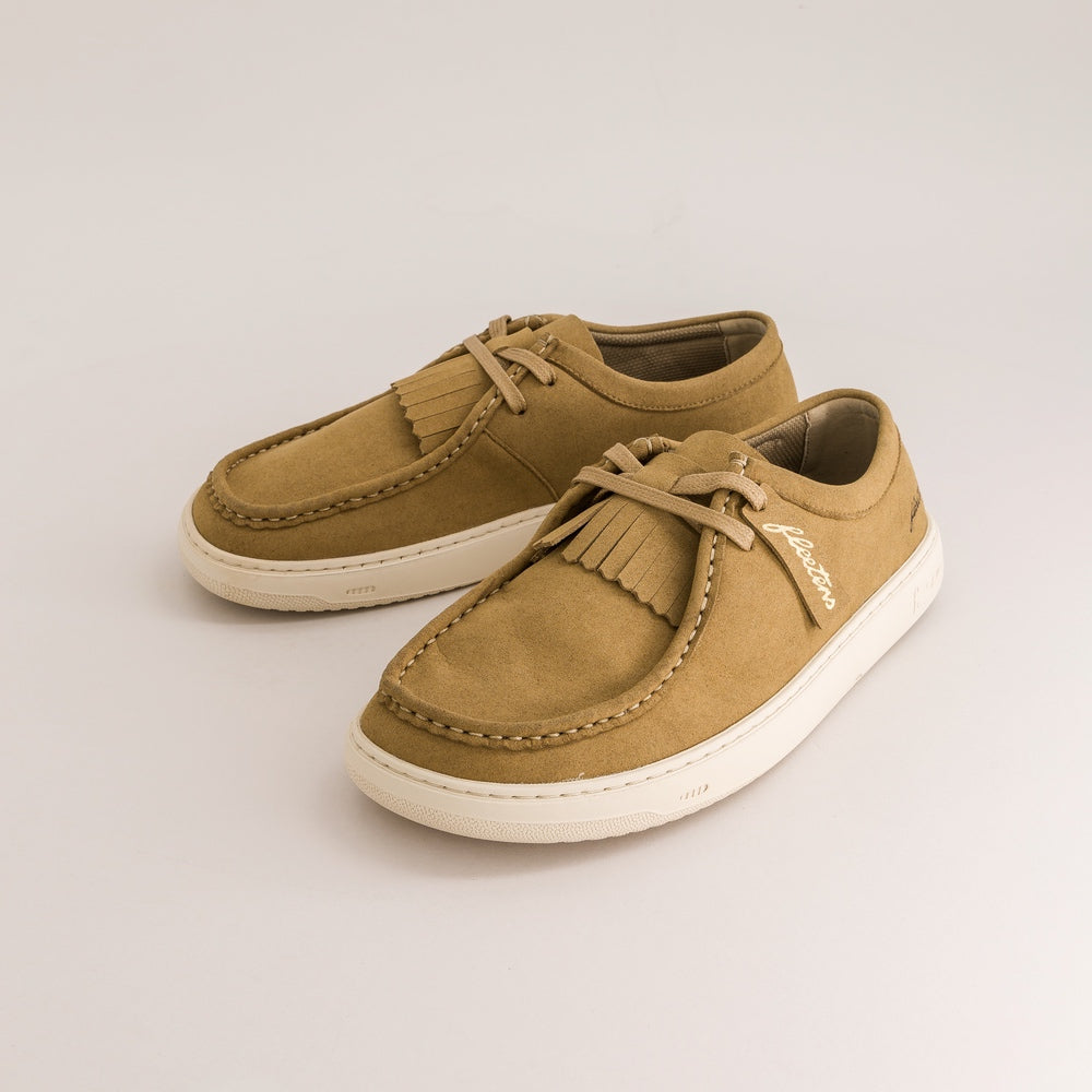 A pair of tan suede derby shoes with fringe tassels on the toe and thick white soles, photographed on a clean light gray studio background.