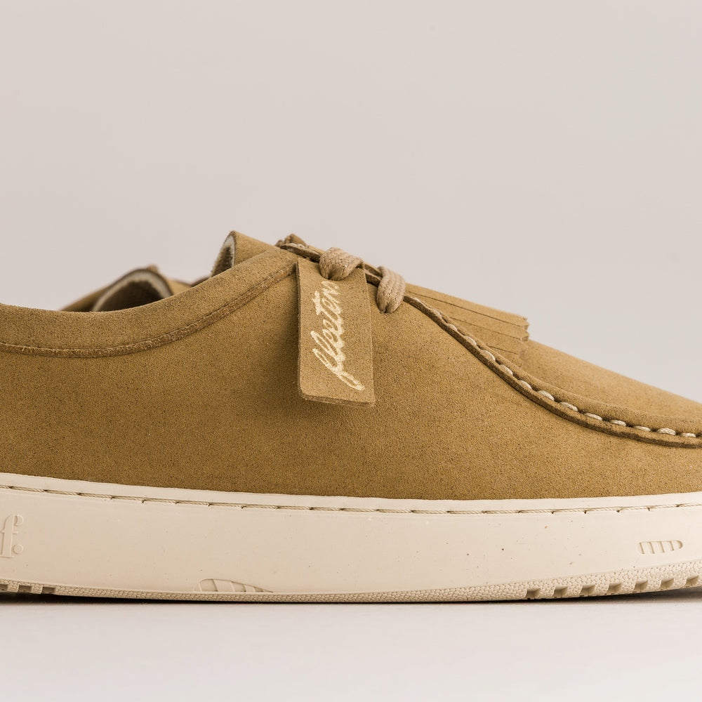 A close-up side view of a tan suede low-top sneaker with a cream-colored sole, featuring a stitched tassel detail and a fabric tag with the brand name 'FasTun'.