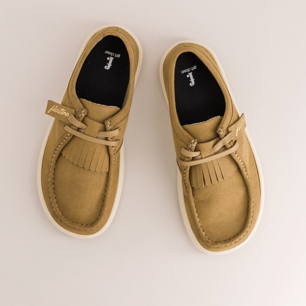 A pair of tan suede moccasin-style slip-on shoes with fringe detailing and white soles, photographed from a top-down angle on a light gray background.
