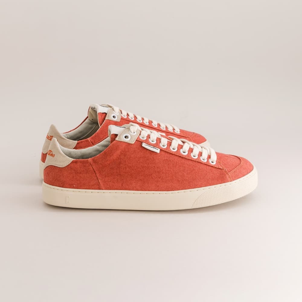 A pair of low-top canvas barefoot sneakers in a warm coral-red color, featuring white laces, white rubber soles, and light beige heel tabs with a small orange logo.