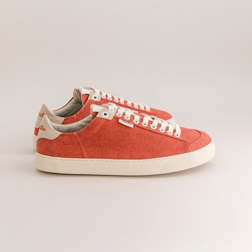 A pair of low-top canvas barefoot sneakers in a warm coral-red color, featuring white laces, white rubber soles, and light beige heel tabs with a small orange logo.