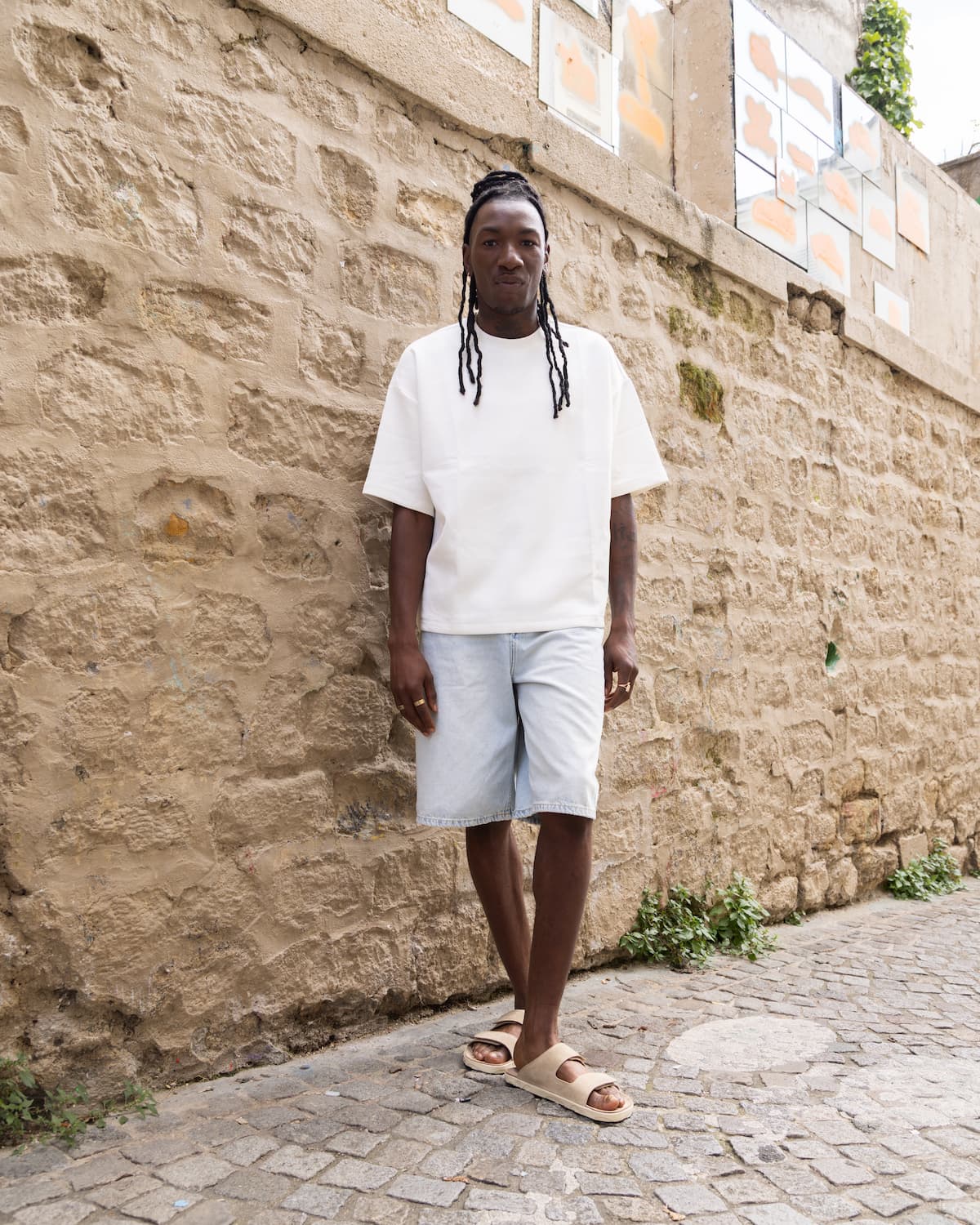 Person wearing a white t-shirt and light shorts standing against a stone wall.