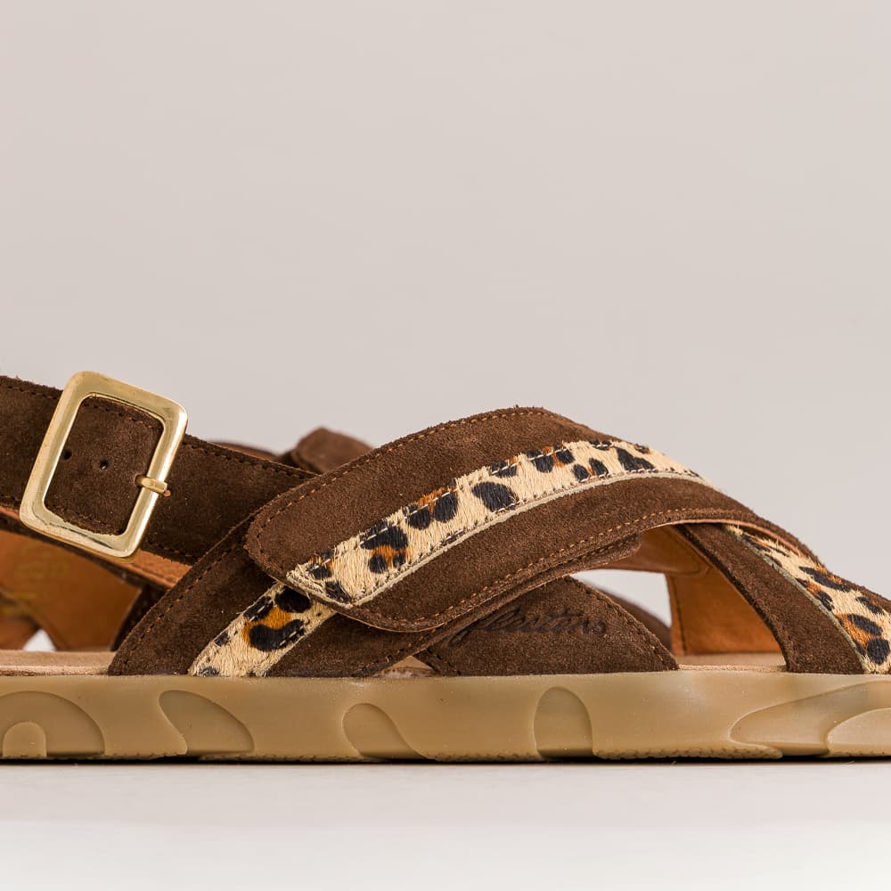 A close-up side view of a brown leather sandal with leopard-print straps and a gold buckle, set against a neutral light gray background.