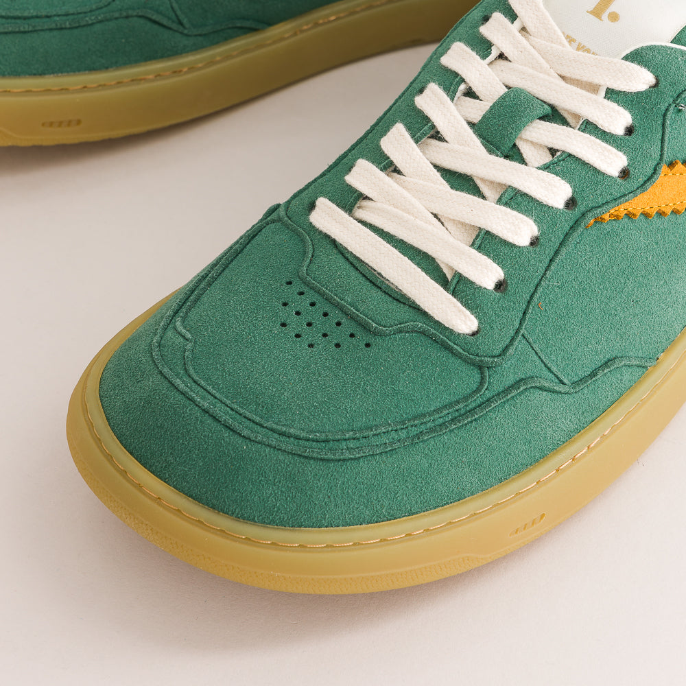 Close-up of a green suede sneaker with white laces, a yellow side accent, and a gum-colored sole on a light background.