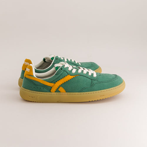 A pair of green suede sneakers with yellow side stripes, white laces, and a gum sole, photographed on a light beige studio background.