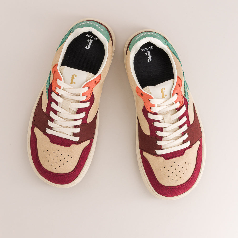 A pair of low-top sneakers shown from a top-down view on a light beige background. The shoes feature a color-blocked design with cream, burgundy, and teal panels, accented with orange laces and a white midsole.