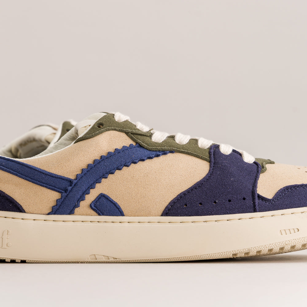 A low-top sneaker shown in a side profile view against a light beige background. The shoe features a beige suede upper with navy blue suede overlays, olive green suede panels, white laces, and a thick cream-colored rubber sole.