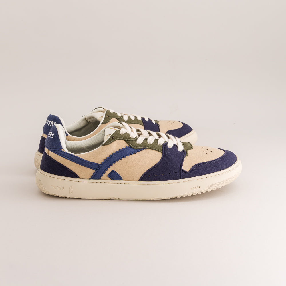 A pair of low-top sneakers in navy blue suede and beige canvas with olive green accents, white laces, and off-white soles, photographed on a light gray studio background.