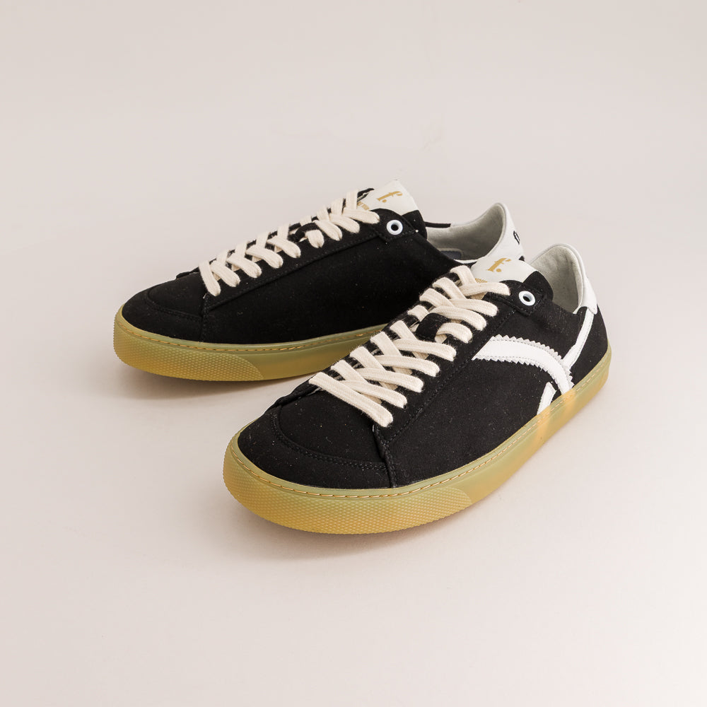 A pair of black canvas low-top sneakers with off-white laces, a white side stripe, and a gum-colored sole, photographed on a clean light beige studio background.