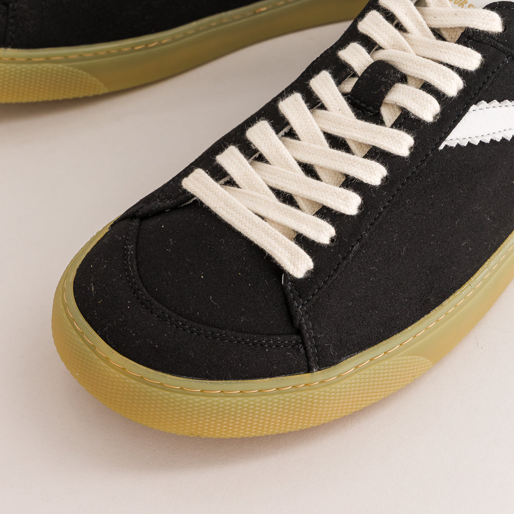Close-up of a black canvas sneaker with off-white laces and a gum rubber sole, shown on a light beige background.
