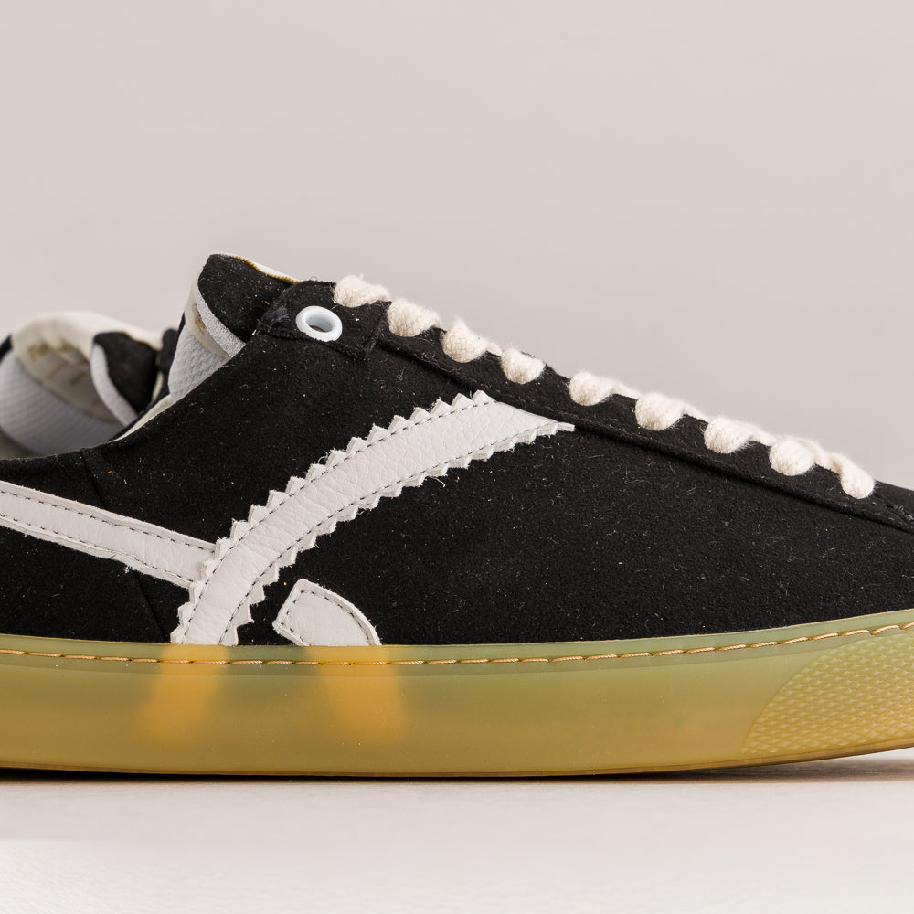A close-up side view of a black suede sneaker with white contrast stitching, white laces, and a translucent yellow gum sole, shown against a neutral light gray background.
