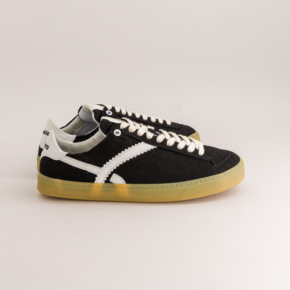 A pair of black suede low-top sneakers with white side stripes, white laces, and translucent yellow gum soles, photographed on a clean light gray studio background.