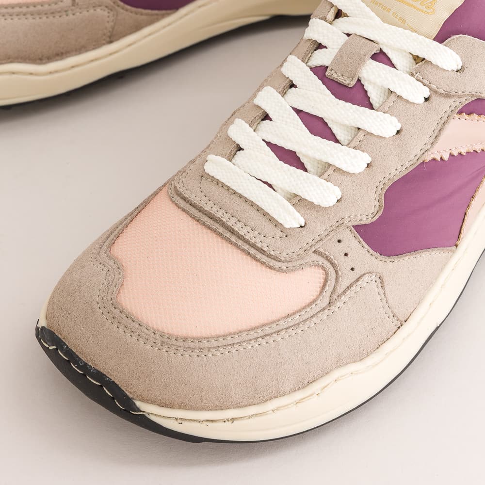 Close-up of a low-top barefoot sneaker showing the toe box, featuring beige suede and mesh panels, a light pink overlay, purple side accents, white laces, and a cream midsole.