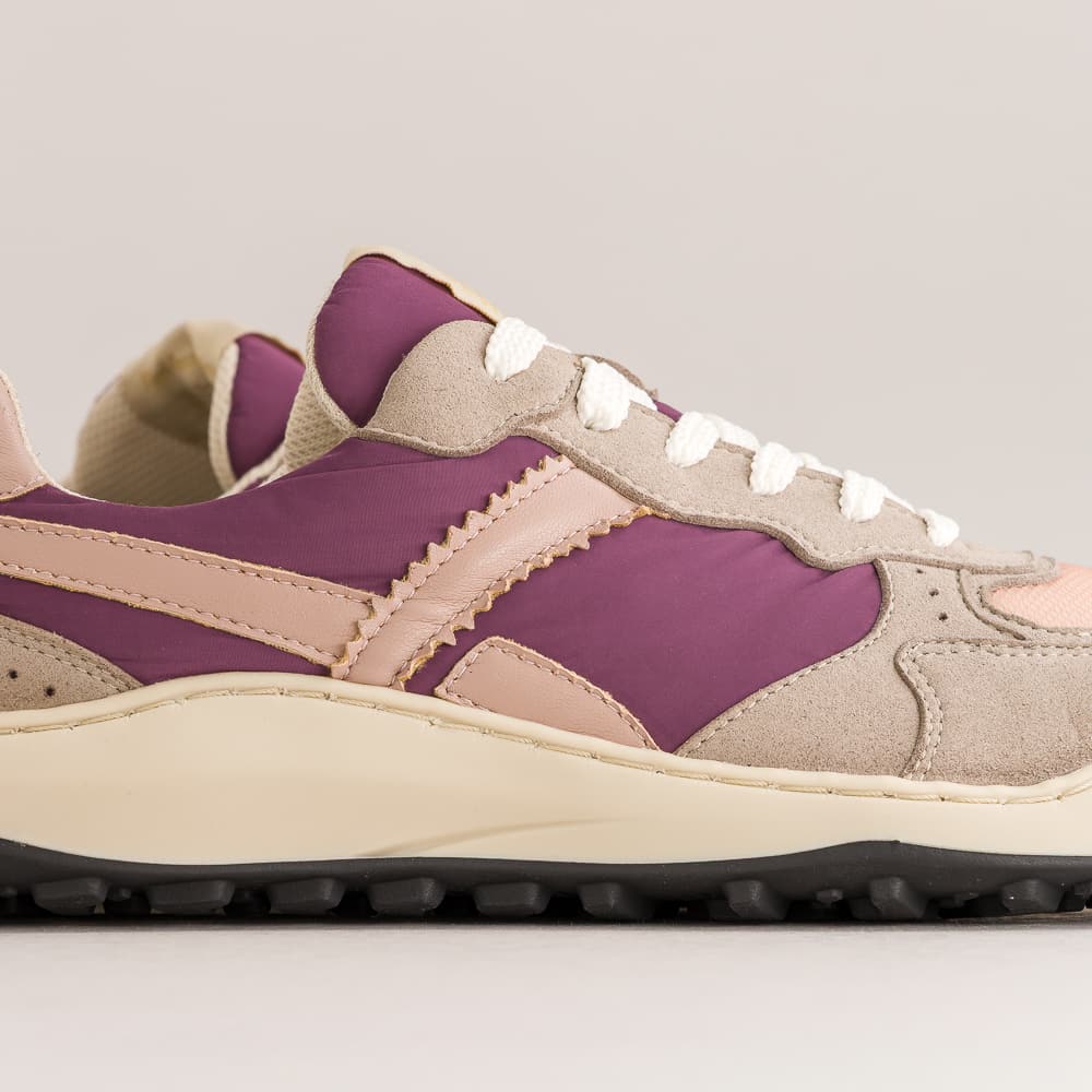 A close-up side view of a low-top barefoot sneaker featuring a color-blocked design with purple panels, beige suede overlays, a light pink leather stripe, white laces, and a cream midsole on a light gray background.