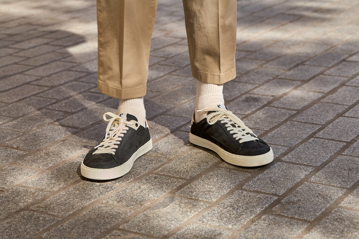 A person wearing black barefoot sneakers with off-white laces and soles, paired with beige trousers and white socks, standing on a gray paved outdoor walkway.