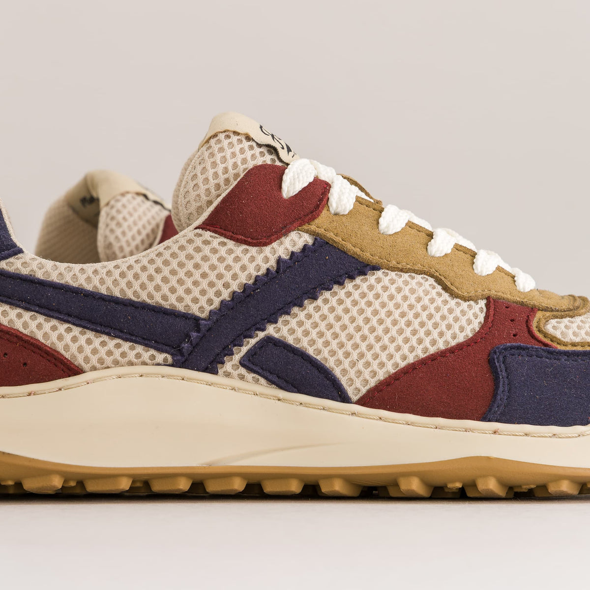 A close-up side profile of a multi-colored retro barefoot sneaker. The shoe features a beige mesh base with navy blue suede overlays, burgundy suede panels, tan leather accents, and white laces, set against a neutral light gray background.