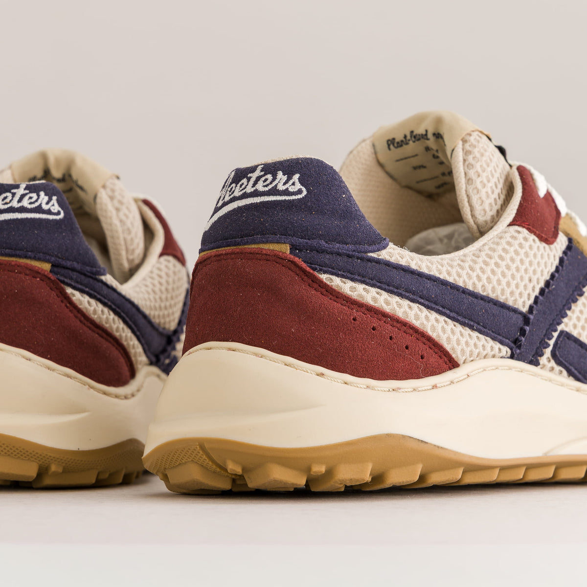 Close-up of the rear half of a pair of barefoot sneakers, showing cream mesh and suede, navy blue overlays, burgundy suede panels, and a cream midsole with a tan outsole.