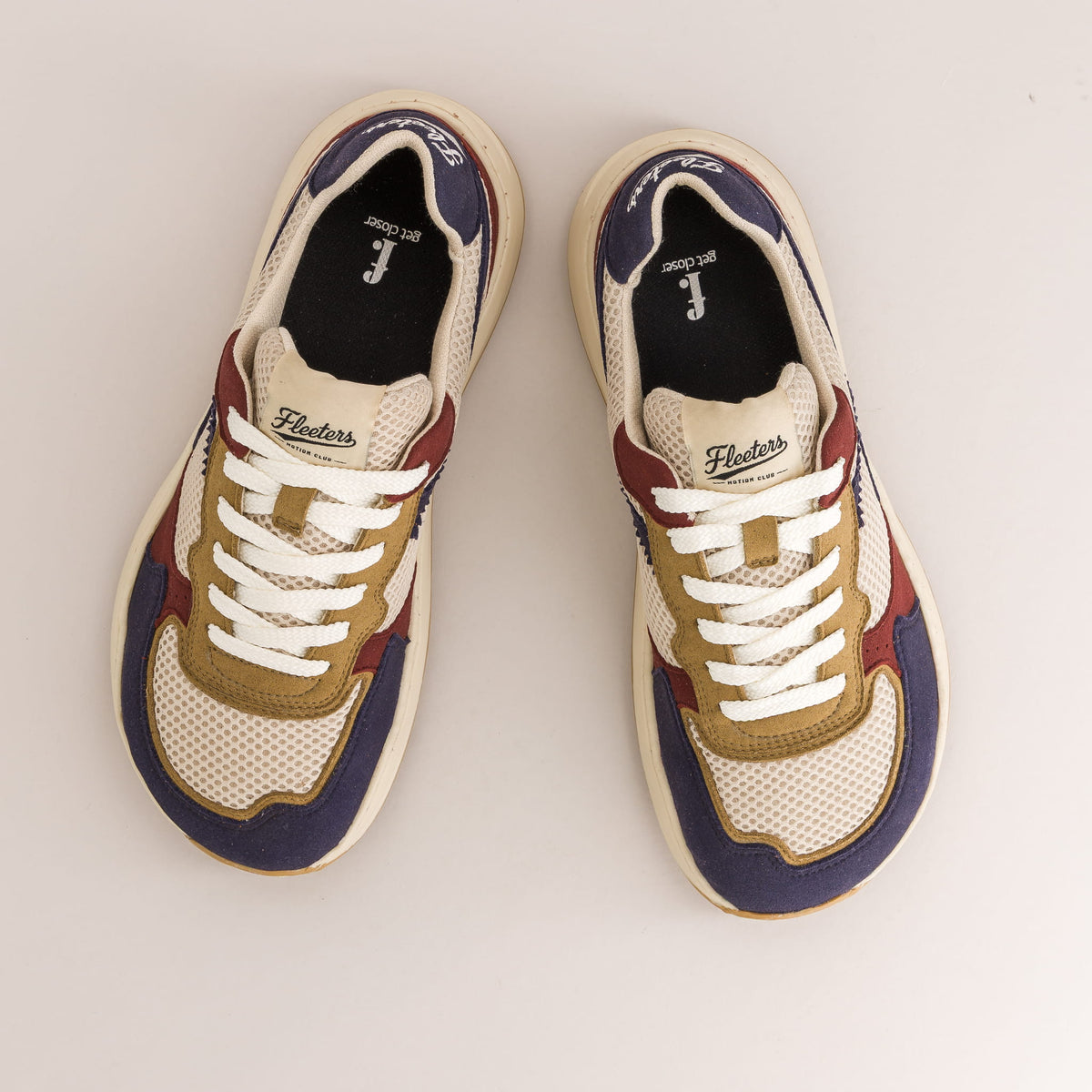 A pair of low-top barefoot sneakers shown from a top-down view on a light beige background. The shoes feature a cream mesh base with navy blue panels, tan overlays, burgundy side accents, and white laces.