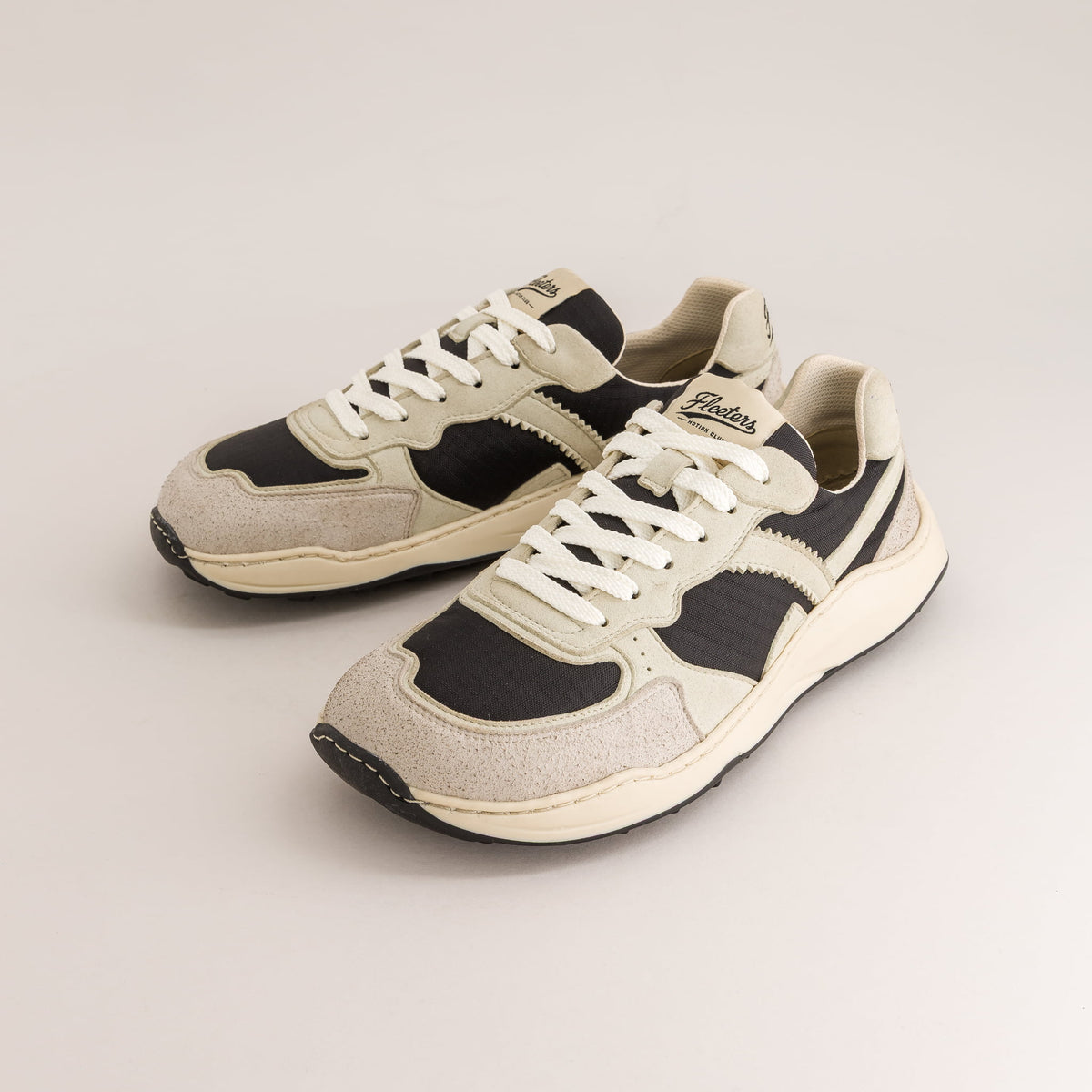 A pair of retro-style low-top barefoot sneakers in beige, black, and off-white, featuring white laces and a chunky cream midsole, photographed on a light beige studio background.