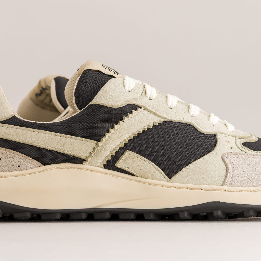 A close-up side profile of a low-top barefoot sneaker in beige and black, featuring a thick cream midsole and black outsole, set against a neutral light gray background.