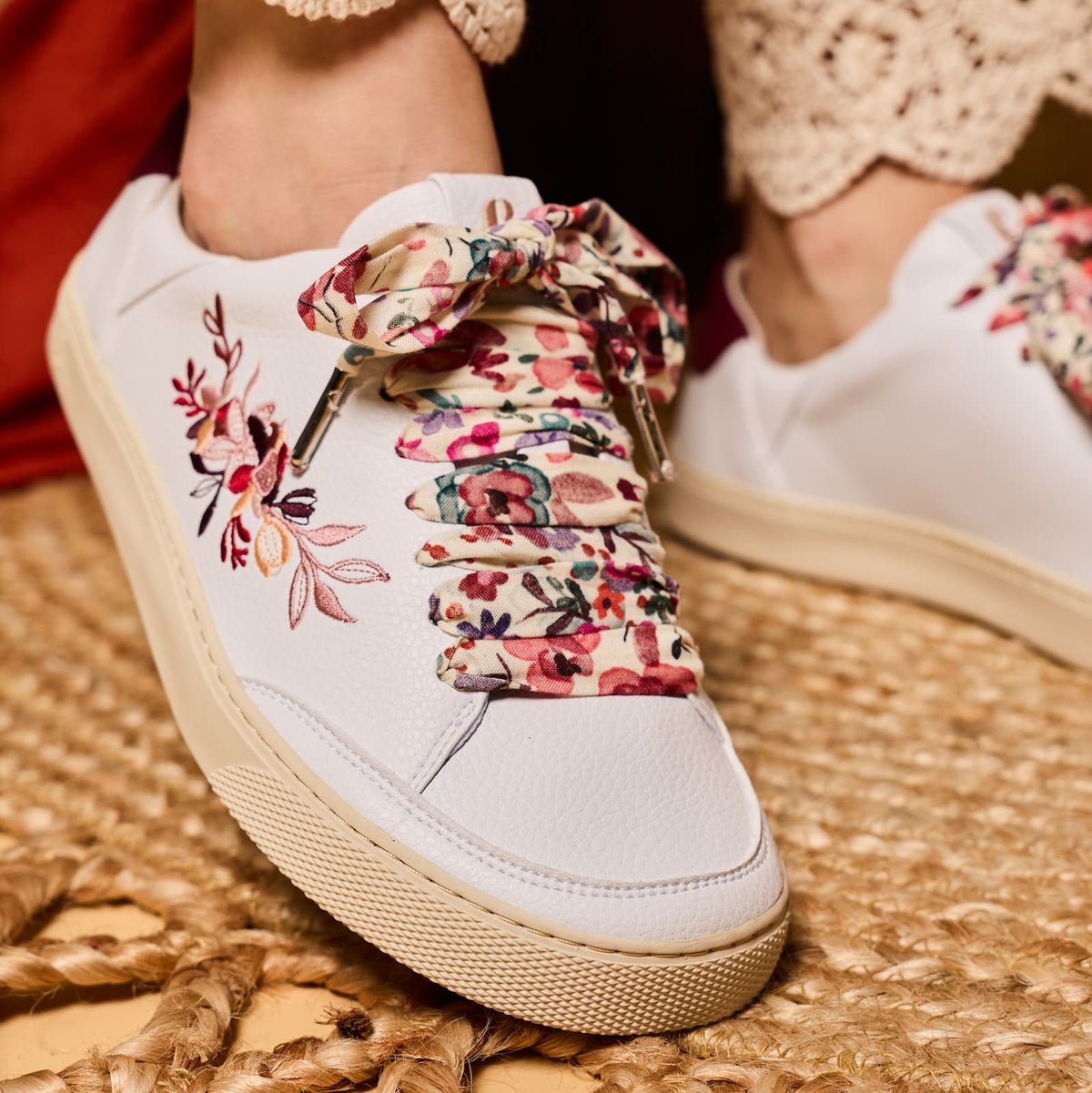 White low-top barefoot sneakers with colorful floral lace-up detailing and floral embroidery on the side, shown being worn with beige lace-trimmed pants on a woven mat.