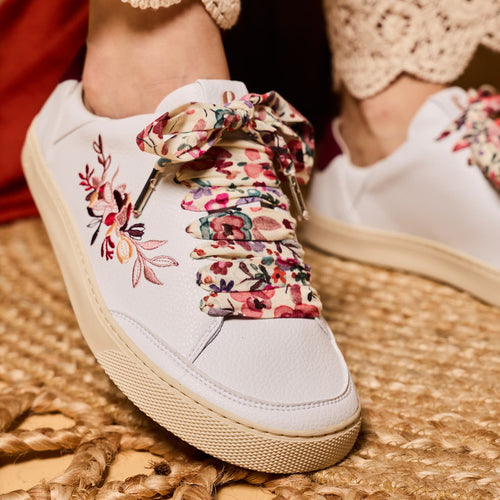 White low-top barefoot sneakers with colorful floral lace-up detailing and floral embroidery on the side, shown being worn with beige lace-trimmed pants on a woven mat.
