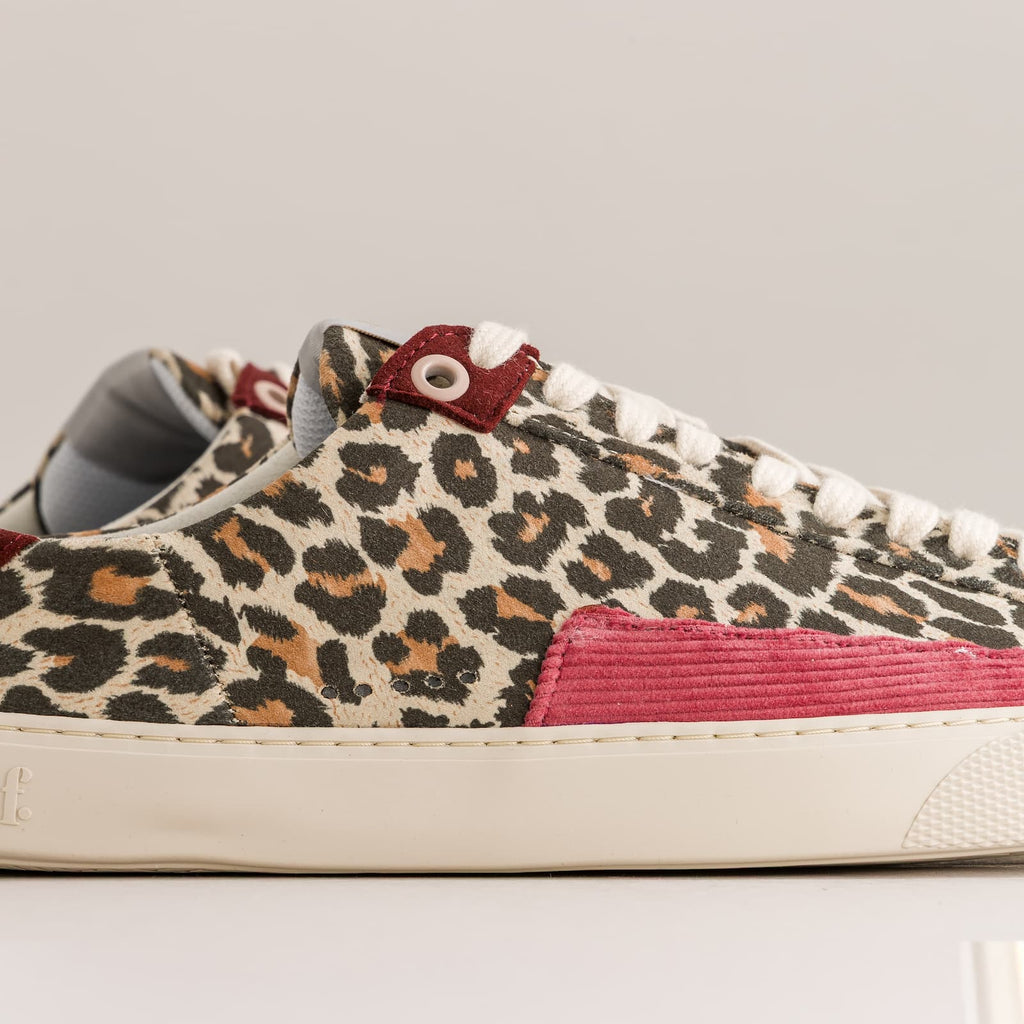 A close-up side view of a low-top barefoot sneaker featuring a beige, brown, and black leopard print canvas upper, a vibrant pink corduroy side panel, white laces, and a cream-colored rubber sole.