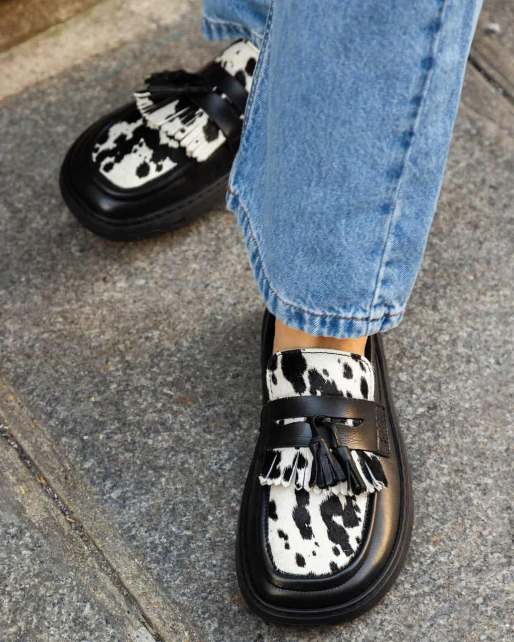 Black and white cow print loafers worn with blue jeans on a pavement.