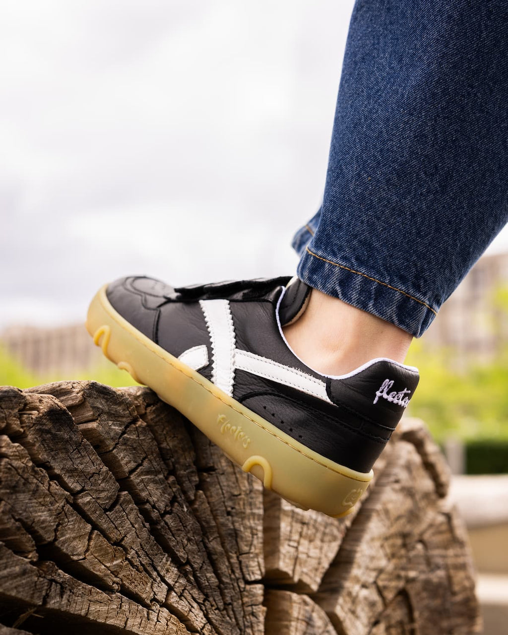 A kid wearing blue jeans stands on a weathered log outdoors, showcasing a black and white barefoot sneaker with a beige sole.