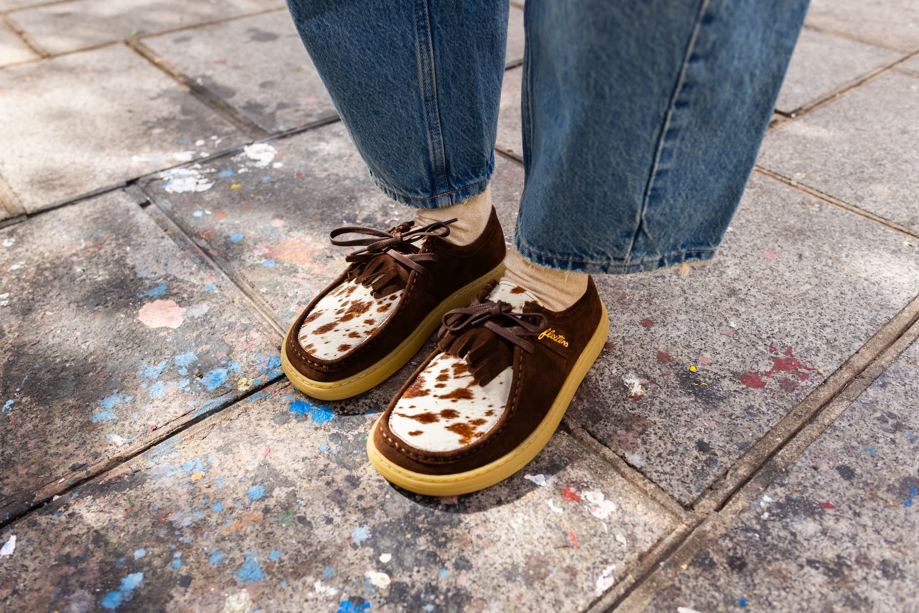 A person wearing brown suede barefoot derby shoes with a white cow-print pattern and dark brown laces, paired with blue jeans, standing on a gray concrete pavement.