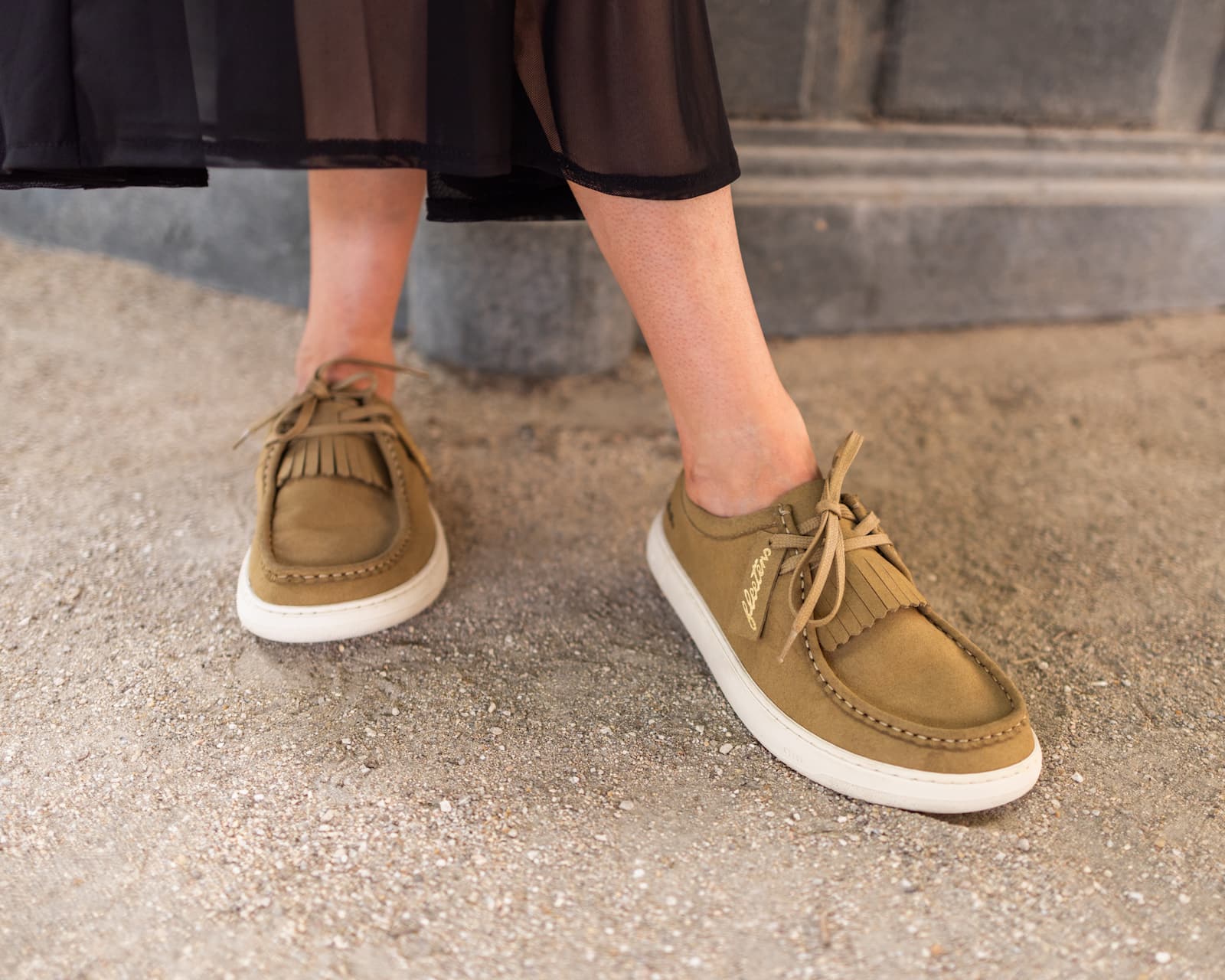 A person wearing tan suede moccasin-style barefoot shoes with fringe and white soles, standing on a gray concrete surface.