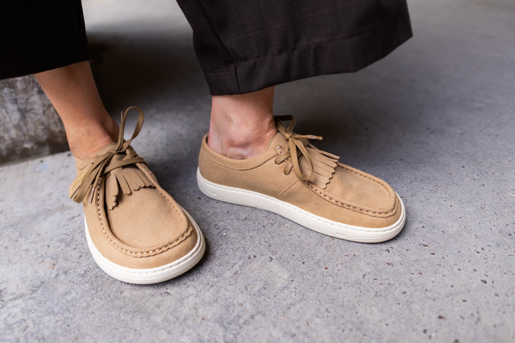 A close-up of a person wearing tan suede barefoot derby shoes with white soles, standing on a gray concrete floor.