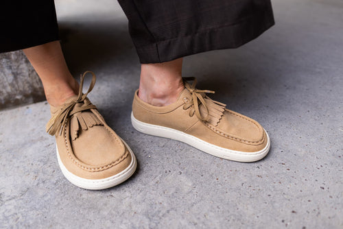 A close-up of a person wearing tan suede barefoot derby shoes with white soles, standing on a gray concrete floor.