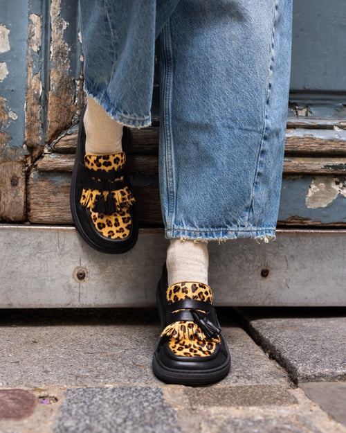 A person wearing leopard-print barefoot loafers with black straps and tassel details, paired with light blue jeans and white socks, sitting on weathered wooden steps.