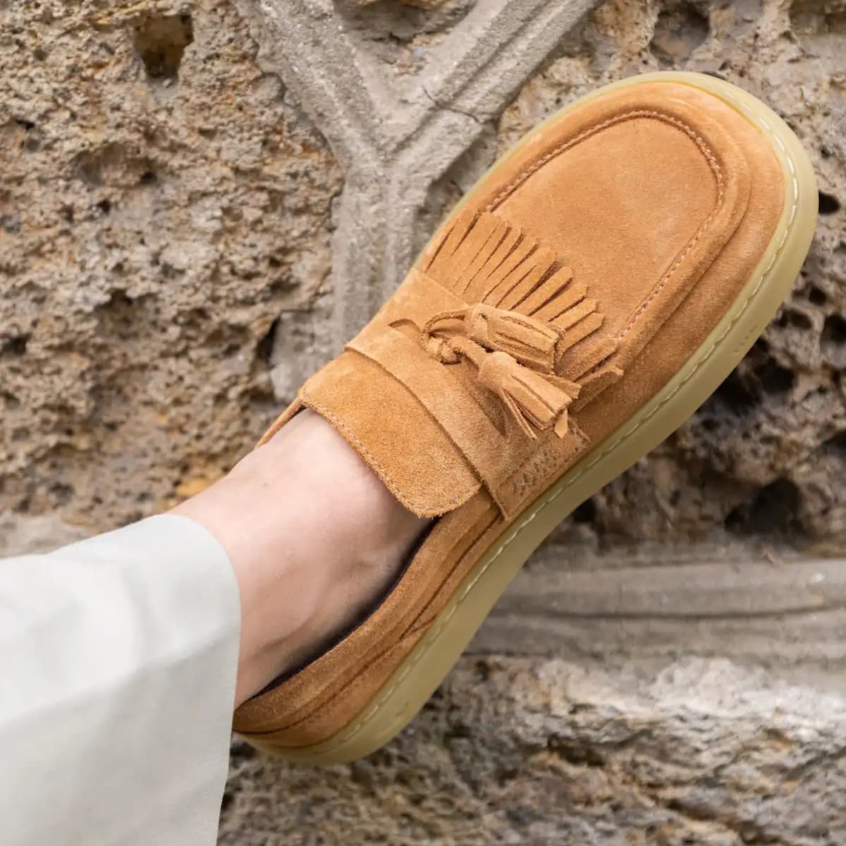 A tan suede tassel loafer barefoot shoe worn on a foot, shown against a textured stone wall background.