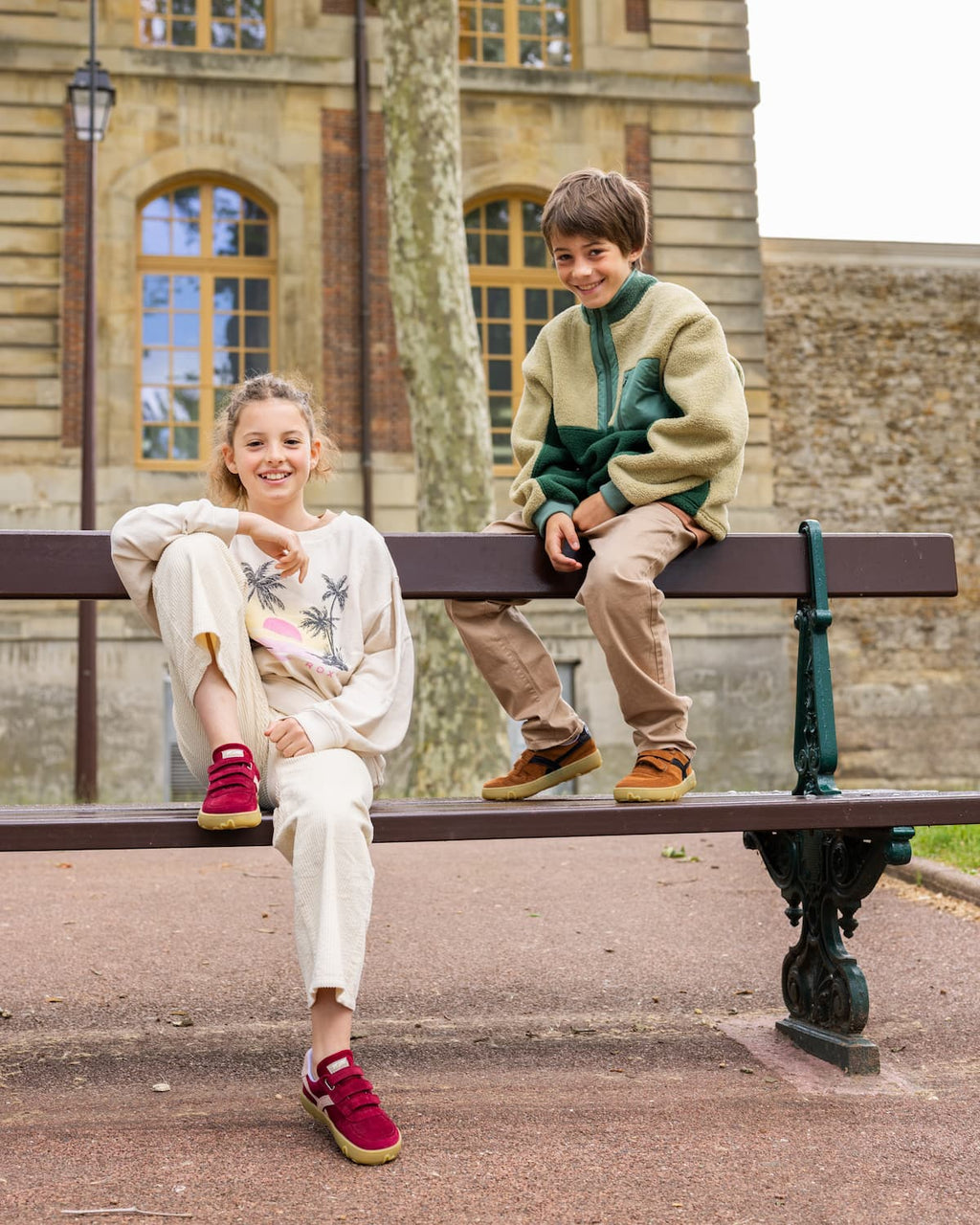 Two children sitting on a park bench in front of a stone building. The child on the left wears a cream sweatshirt with a tropical print, cream pants, and burgundy barefoot sneakers.