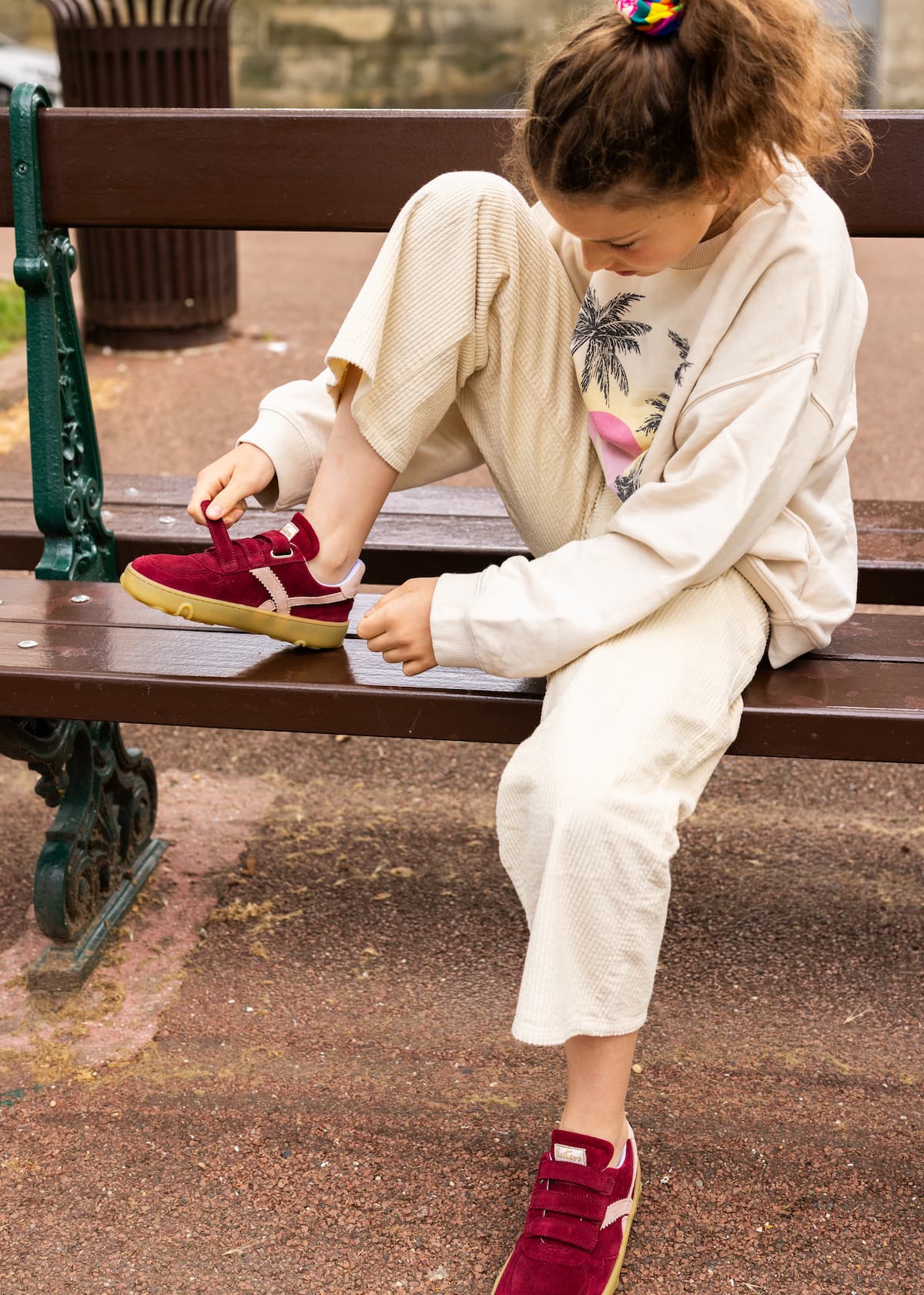 A young girl sitting on a wooden park bench, tying a burgundy barefoot sneaker with a yellow sole while wearing a cream-colored knit lounge set.