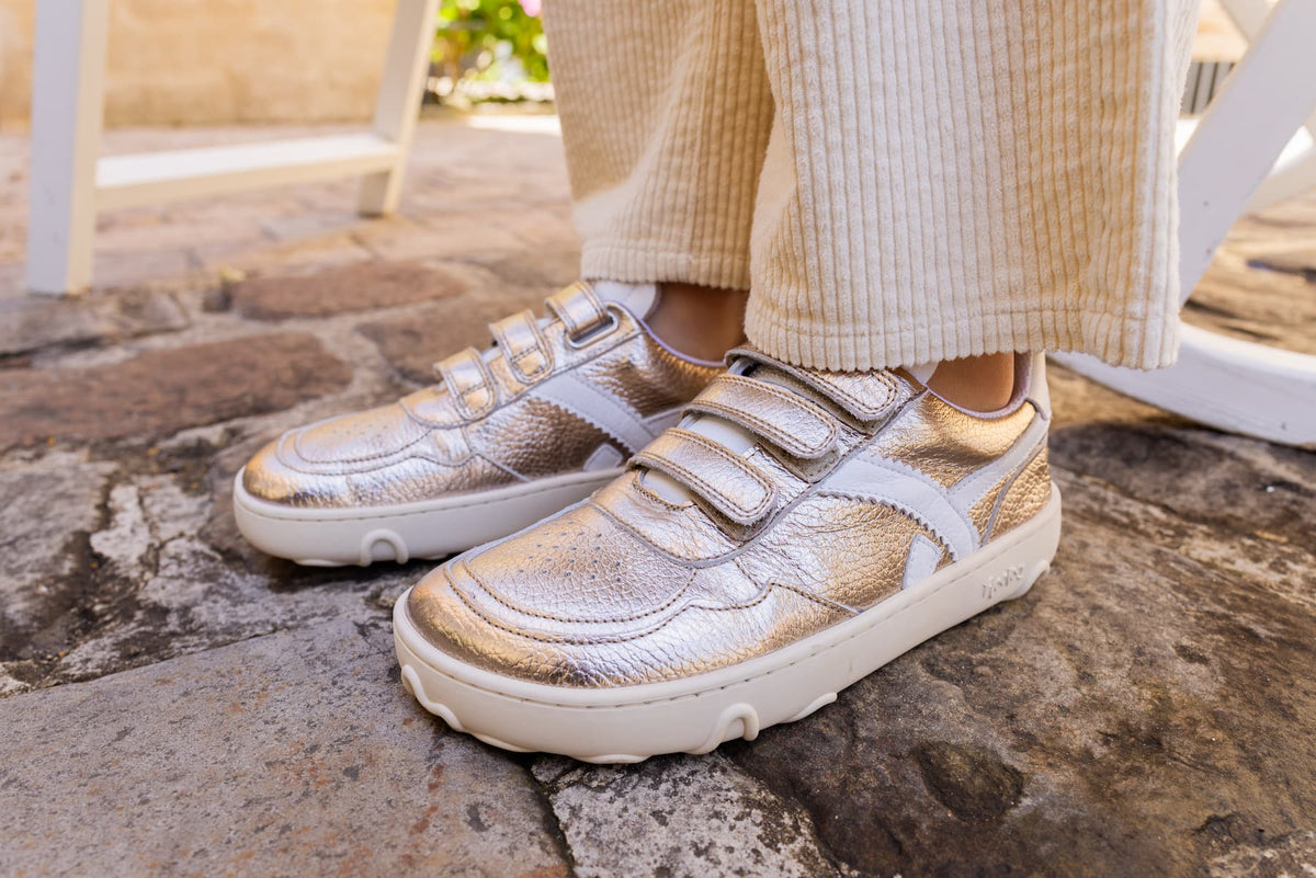 A close-up of a kid wearing metallic gold and white low-top barefoot sneakers with double hook-and-loop straps, standing on a stone patio.